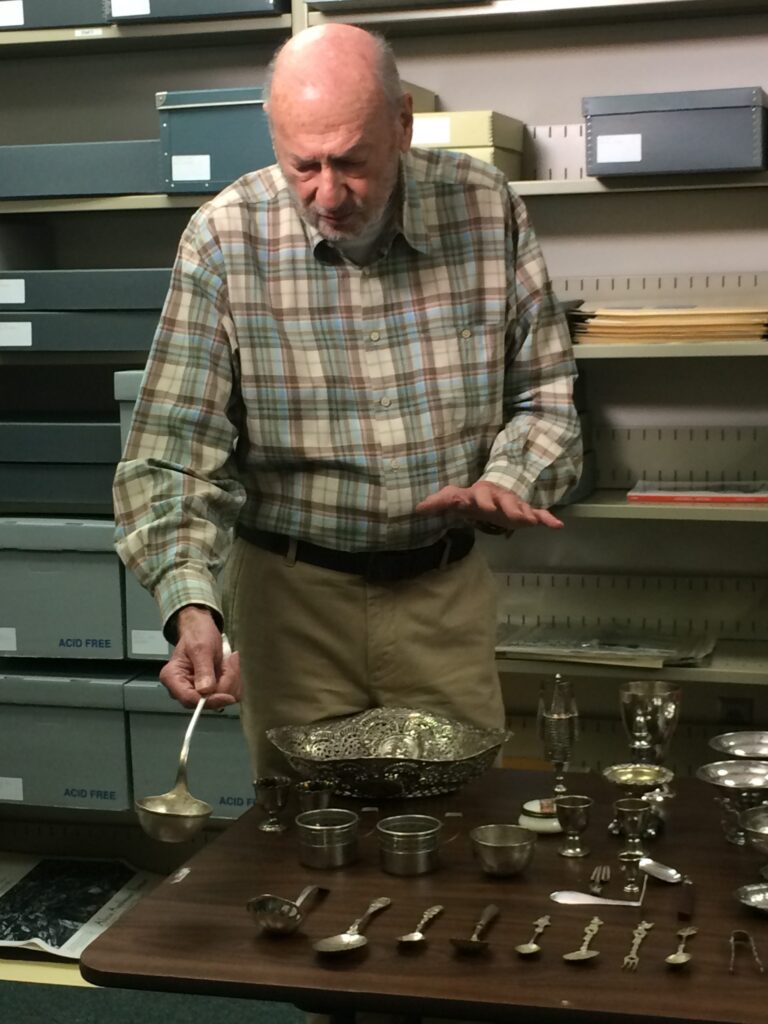 Holocaust Survivor Werner Knurr standing with his family’s collection of sabbath items and dinnerware.